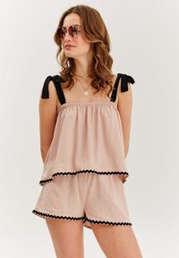 Žena v svetloružovom bezrukávovom topi a kraťasoch s čiernym lemovaním, čiernymi ramennými viazaniami, okrúhlymi slnečnými okuliarmi a dvoma náhrdelníkmi, pozerajúca sa do strany.