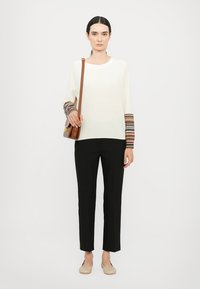 Top bianco a maniche lunghe con polsini a strisce multicolori, pantaloni neri su misura e scarpe beige senza lacci, accessoriate con una borsa a tracolla marrone.