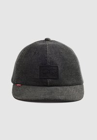 Gorra de pana gris con una superficie texturizada, visera plana y un parche de cuero negro que muestra el logotipo de la marca en la parte frontal.