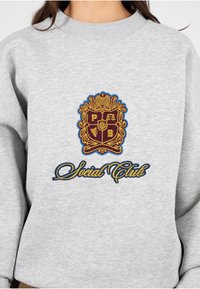 Grijze sweatshirt met een groot geborduurd wapen in bordeauxrood en goud, met de tekst "Social Club" in een sierlijk lettertype.