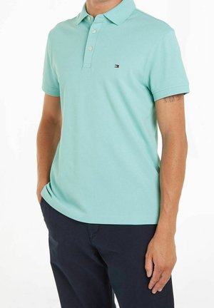 Homme portant un polo bleu clair à manches courtes avec trois boutons et un pantalon bleu marine, debout les mains dans les poches.