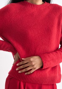 Pull rouge texturé avec un col rond et des poignets côtelés. Il présente une coupe décontractée et est accompagné de pantalons unis à proximité.