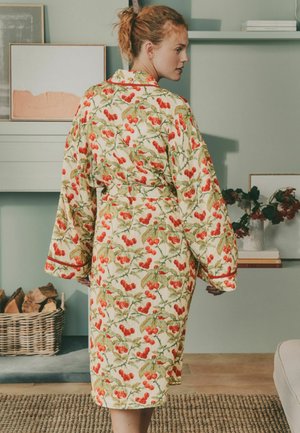 Woman wearing a cherry-patterned robe with wide sleeves stands indoors near a basket of firewood and a shelf with books and flowers.