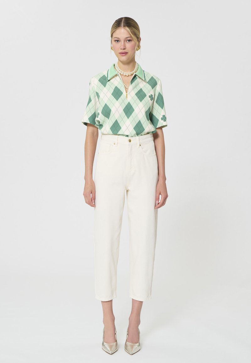 Femme portant un polo à manches courtes à motif losanges vert et blanc, un pantalon blanc court taille haute, un collier de perles et des talons métalliques pointus.
