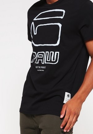 Osoba nosząca czarną koszulkę G-Star RAW z dużą białą grafiką i tekstem na przodzie, w połączeniu z ciemnozielonymi spodniami.