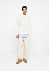Pulover alb din tricot cu guler rotund, suprapus peste o cămașă în dungi albastre deschise, asortat cu pantaloni bej de dimensiuni mari și teniși maronii.