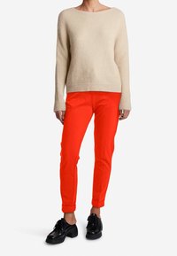 Beige gestrickter Pullover mit weitem Ausschnitt, kombiniert mit leuchtend roten Hosen. Schwarze Schnürschuhe mit einem klobigen Absatz runden das Outfit ab.