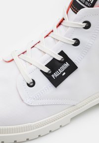 Sneaker alta in tela bianca con accenti neri e arancioni, dotata di etichetta nera, lacci bianchi e suola in gomma con grip testurizzato.