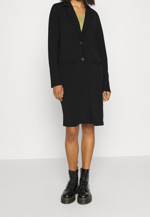 Kvinde iført en knælængde sort knaplukket frakke over en grøn top, kombineret med sorte snørestøvler med plateau mod en ensfarvet baggrund.