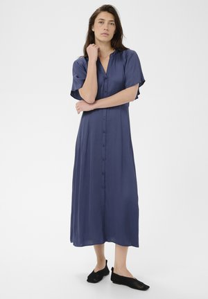 Ženska stoji z eno roko ob bradi, oblečena v modro midi obleko z gumbi in črne mrežaste copate brez zavezovanja na belem ozadju.