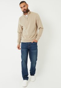 Pull en beige à zip quart en matériau doux, associé à un jean en denim bleu et des baskets blanches. Design simple avec des poignets côtelés.