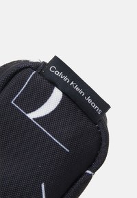 Juodas audinio maišelis, puikuojantis geometriniu baltu raštu, šone esančia etikete "Calvin Klein Jeans". Lygus paviršius.