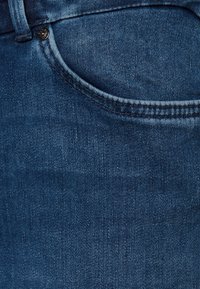 Dunkelblaue Denim-Stoff zeigt feine Textur, Taschennaht und einen Metallknopf-Akzent, mit einer glatten Oberfläche und subtilen Verwaschungen.