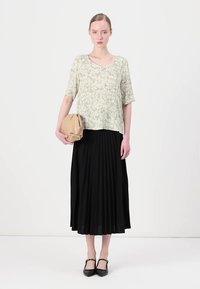 Lys grønn blomsterbluse med korte ermer, sammen med et langt svart plissert skjørt. Holder en beige clutch-veske. Svart sko med stropper.