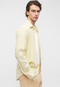 Κίτρινο ριγέ πουκάμισο με κουμπιά, άνετη εφαρμογή και απαλό ύφασμα, φορεμένο πάνω από λευκή μπλούζα. Το μοντέλο φοράει ανοιχτό μπεζ παντελόνι.