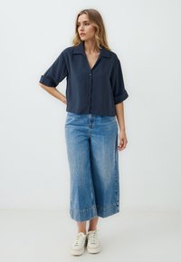Camicia blu navy con bottoni frontali, colletto e maniche arrotolate, abbinata a pantaloni di denim a gamba larga azzurri e sneakers bianche.