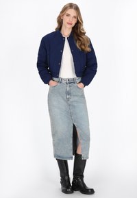 Jachetă bomber matlasată de culoare bleumarin cu nasturi tip snap, top alb, fustă din denim de culoare albastru deschis cu fente în față și cizme negre până la genunchi.