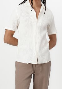 Koszulka z krótkim rękawem w kolorze białym, wykonana z fakturowanej tkaniny, z zapięciem na guziki, kołnierzykiem i prostym dołem. Noszona z beżowymi spodniami.