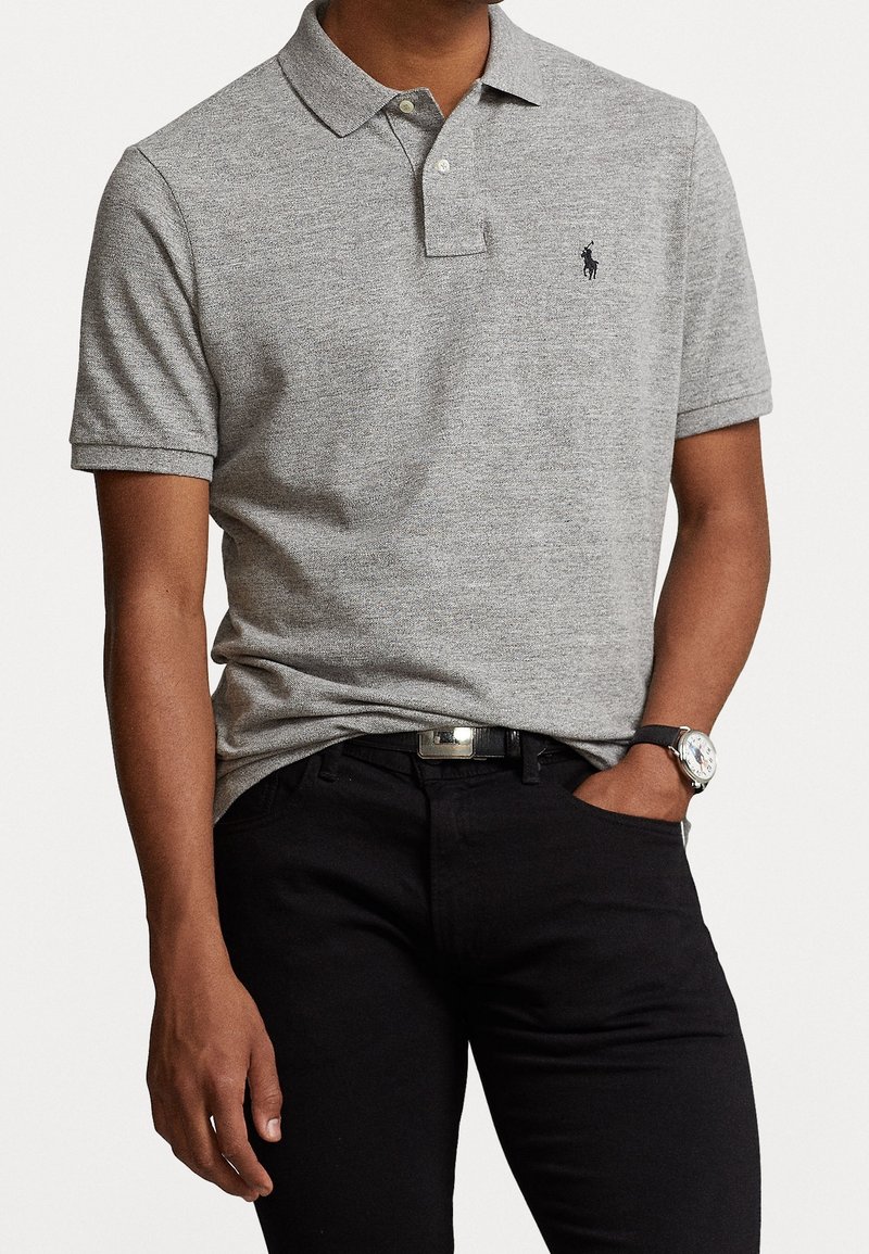 Polo gris à manches courtes, avec deux boutons au col et un logo brodé sur la poitrine gauche, associé à un jean noir et une montre.