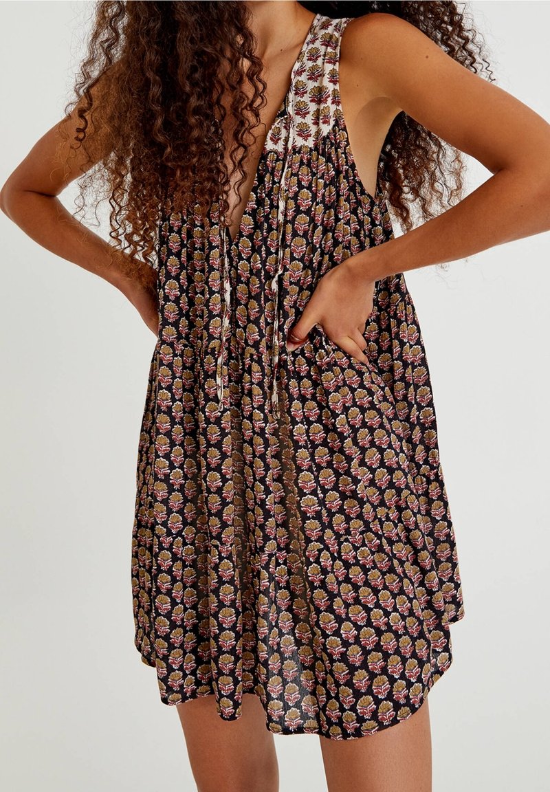 Robe sans manches à motifs floraux roses et jaunes sur fond noir portée par une personne aux longs cheveux bouclés, les mains posées sur les hanches.