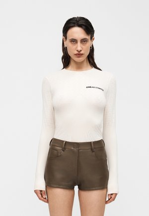 Γυναίκα με σκούρα μαλλιά φοράει λευκή μπλούζα με μακριά μανίκια και κείμενο «SINÉAD O'DWYER» και καφέ σκούρα δερμάτινα σορτς με ψηλή μέση, στέκεται μπροστά σε λευκό φόντο.