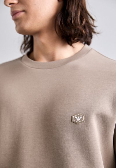 Beige sweatshirt with a crew neck, soft texture, and a hexagonal logo patch on the left side. Simple design, no patterns or noticeable accents.