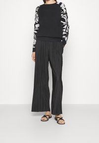 Svart tröja med mönstrade grå ärmar, kombinerad med veckade svarta vida byxor och svarta sandaler med en rosettdetalj.