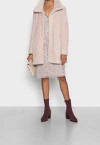 Abrigo de pelo sintético rosa claro con cuello de pico y seis botones, llevado sobre un vestido beige con capas, adornos plateados y botas de tobillo burdeos.