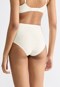 Vrouw in hooggezette witte onderbroek en bijpassende beha, van achteren zichtbaar tegen een effen lichte achtergrond.