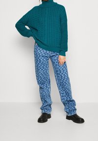 Teal neulepusero, jossa on kauluri ja tekstuurisia kuvioita, yhdistettynä sinisiin kuvioituihin farkkuihin ja mustiin nilkkasuksiin.