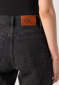 Asmuo vilkintis juodais džinsais su matomu rudu Calvin Klein odiniu logotipo pažymėjimu ant nugarinės juostos, ir juoda palaidine įsikišta.