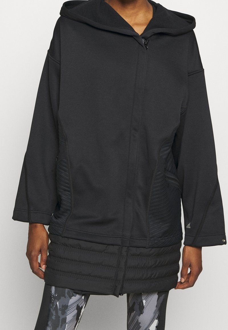 Personne portant une veste de sport noire à capuche et manches longues avec des détails matelassés sur le bas devant et des leggings noirs et blancs à motifs.