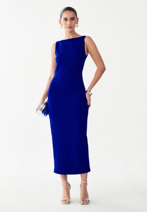 Vrouw draagt een mouwloze midi-jurk in koningsblauw met zilveren hakken, houdt een bijpassende blauwe clutch vast, staand tegen een witte achtergrond.