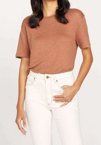 Žena nosící hnědočervenou krátkou košili zastrčenou do bílých zvonových džín, s jednou rukou v kapse a druhou uvolněnou.