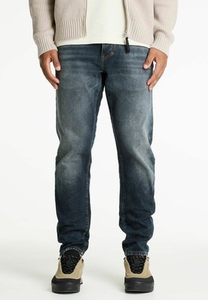 Homme portant un jean bleu délavé, un pull beige, une chemise blanche, et des baskets beige et noires, se tenant devant un fond blanc.