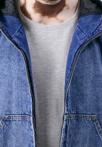 Džínová bunda s modrým vnějškem, šedou podšívkou a dvěma předními kapsami, přehozena přes světle šedé tričko. Viditelný zipový uzávěr.