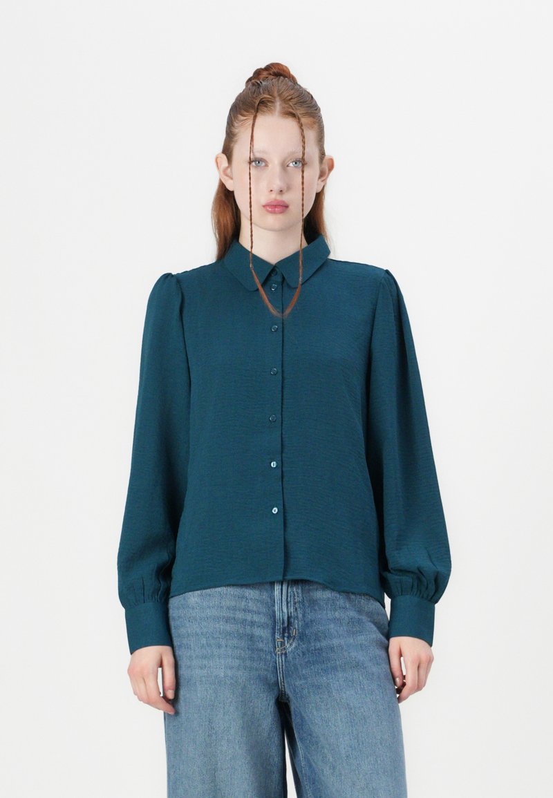 Knoophemd in diep teal met een gestructureerde stof, lange mouwen en een klassieke kraag. Heeft opgevulde schouders en bijpassende knoopdetails.