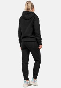 Felpa con cappuccio nera e pantaloni abbinati in tessuto morbido, caratterizzati da una vestibilità rilassata e polsini elastici. Indossati con scarpe da ginnastica di colore chiaro.