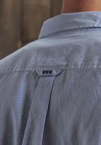 Camisa a rayas azul y blanca con collar abotonado, que presenta una pequeña etiqueta de logo en la parte posterior cerca del cuello. Textura suave.