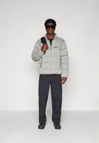 Pelēka pufveida jaka ar priekšējo rāvējslēdzēju un divām sānu kabatām. Kombinēta ar melnām biksēm un kurpēm. Modelis valkā saulesbrilles un tur siksniņu.