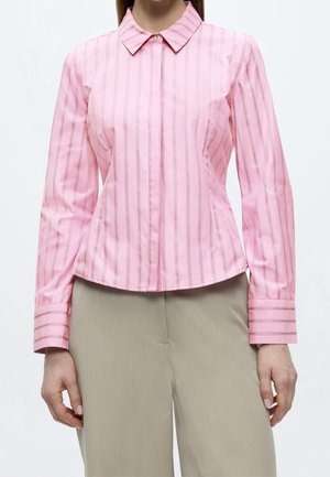 Vrouw draagt een aansluitend roze langarmshirt met verticale strepen en beige broek, staand tegen een eenvoudige lichte achtergrond.