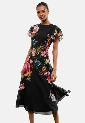 Frau trägt ein schwarzes Midikleid mit bunten Blumen- und Schmetterlingsmustern, flatternden Ärmeln und einem fließenden Rock, posiert vor weißem Hintergrund.