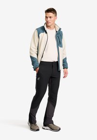 Herrernas vandringsoutfit som innehåller en tvåfärgad softshelljacka i kräm och teal, slim-fit svarta byxor och grå vandraskor. Tydlig logotyp synlig.