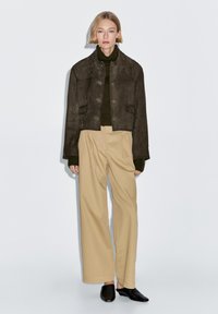 Bruine cropped jas van suède met grote knopen, gecombineerd met een donkergroene coltrui, beige wijde broekspijpen en zwarte instapschoenen.