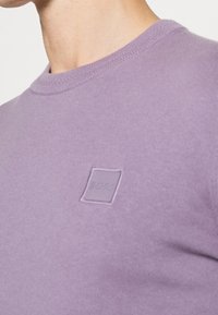 Pull col rond violet en tissu doux avec un patch logo subtil au centre et des détails côtelés au niveau du col.