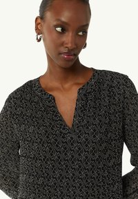 Schwarze Bluse mit V-Ausschnitt, die ein geometrisches weißes Muster aufweist. Strukturierter Stoff, lange Ärmel und Faltendetails an der Brust.