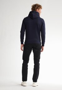 Marineblauwe hoodie met lange mouwen, ribbelboorden en zoom, gedragen met zwarte jeans en witte sneakers. Achteraanzicht, gladde stoftextuur.