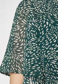 Vihreä puoliksi läpinäkyvä paita, jossa on valkoinen pisaramäinen kuvio, lyhyet liehuvat hihat ja pehmeä, virtaava rakenne.