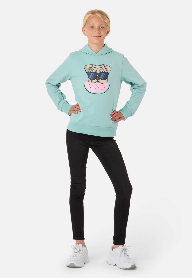 Felpa azzurra chiara con un'illustrazione di un carlino che indossa occhiali da sole e una fetta di anguria, abbinata a jeans skinny neri e sneakers bianche.