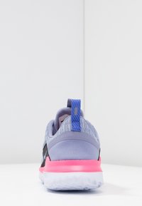 Zapatilla deportiva con parte superior morada, diseño texturizado, acentos negros y suela rosa. Presenta una pestaña azul en el talón para facilitar el calzado.